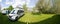 Panorama view of two camper vans parked in an idyllic green farm field with blossoming trees in spring