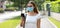 Panoramic banner view of young woman with medical mask messaging on mobile phone in city street. Worried serious girl having smart