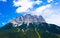 Panoramic drone view from rugged Zugspitze under blue sunny sky with cumulus clouds. Green valley at the foot of majestic alpine m