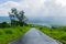 Panoramic landscape view of a rural road with lush green foliage on both sides on a monsoon day, going downhill to Tapola village