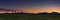 Panoramic shot of a grassy field surrounded by hill silhouettes during a breathtaking sunset