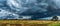 A panoramic shot of a vast green field under the looming presence of dramatic storm clouds, highlighting nature's