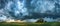 A panoramic shot of a vast green field under the looming presence of dramatic storm clouds, highlighting nature's