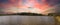 A panoramic shot of a vast rippling lake surrounded by lush green trees, grass and plants with powerful red clouds at sunset