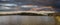 A panoramic shot of a vast rippling lake surrounded by lush green trees with powerful red and yellow clouds with blue sky