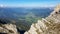 A panoramic view from an Alpine top on a vast valley. There are sharp mountains and high peaks around. Alpine slopes