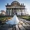 Panoramic view of a fairytale newlywed couple hugging and kissing in front of old baroque ghotic church with columns at sunset
