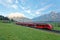 Panoramic view of a train traveling on green fields with Mountain Zugspitze in background on a beautiful sunny day in Lermoos,