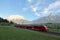 Panoramic view of a train traveling on green fields with Mountain Zugspitze in background on a beautiful sunny day in Lermoos