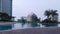 Panoramic view of a wide pool deck in a windy day with palm trees and plants around modern buildings