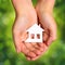 Paper House in Female Hands over Nature Green Sunny Background.