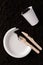 Paper plate and glass, wooden fork and knife on soil close-up, top view. Compostable or biodegradable dinnerware concept