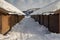 Parallel brown multiple garage buildings, snow on the ground, mountains and white clouds on blue sky