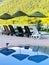 Parasols and sunbeds are reflected on the water of a swimming pool. View of a hill with blooming mimosa trees.