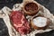 Parchment paper with raw meat fillet near wooden bowls with salt and pepper on black marble surface.