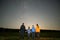 Parents and their kids standing in night field observing dark sky with many bright stars