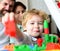 Parents watch son with funny face making toy brick constructions.