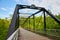 Park with green forest wall behind large dark metal bridge, black beams, blue sky