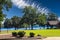 A park at the lake with green grass, a brown wooden pergola, lush green trees with people relaxing on the banks of the lake