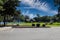 A park at the lake with green grass, a brown wooden pergola, lush green trees with people relaxing on the banks of the lake