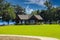 A park at the lake with green grass, a brown wooden pergola, lush green trees with people relaxing on the banks of the lake