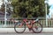 Parked bike in summer in city, red black steering wheel and chair with wheels. Background tree fence pillars with lamps