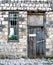 Part of an abandoned house with crumbling walls, broken glass, boarded up dilapidated door and an old pavement