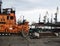 Part of a fire orange towing vessel against the background of a tanker and a port crane and a train with coal