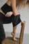 Partial shot of curly woman in dark top and jeans posing near wooden chair on grey background