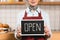 Partial view of barista holding signboard with open inscription in coffee shop.