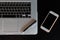 Partial view of a silver keyboard of a laptop with a smartphone and a pencil on a dark wood desk