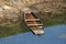 Partially submerged old wooden river boat next to muddy river bank surrounded with floating grass and clear water