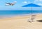 Passenger airplane landing above tropical beach with white wooden beach chair and blue parasol