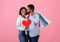 Passionate black man holding shopping bags, kissing his girlfriend with red heart on pink studio background