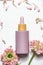 Pastel purple cosmetic bottle with pipette on white background with pink flowers , top view. Modern beauty product for skin care