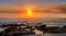 Pastel shades of yellow, red and blue reflecting on the ocean in the setting sun with rocks on the beach.