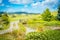 Pastoral rural landscape with gravel road winding past lily pond towards lush green rolling hills.