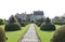 A path edged with with rounded topiary bushes with a conical top leads up to an old English Country House