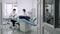 Patient sits down on blue dentist chair, assistant of dentist help to him.