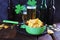Patrick Day, foamy beer in glass mugs and a bottle, paprika chips, gold coins, on a red wooden table, a green shamrock
