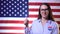 A patriotic woman with a voit sign on her chest stands against the backdrop of the American flag, smiles and shows a