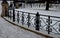 Paved granite cube path in the park. lawns covered with snow and fenced with a formal gray metal fence. Separating the pavement fr