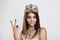 Peace. Beauty queen girl with crown, princess on white gray background, shows victory two 2 sign gesture with fingers hand and