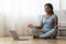 Peaceful african lady meditating in front of laptop at home, practicing yoga