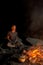 Peaceful mature man, tourist sitting with eyes closed near campfire and cooking dinner outdoors in forest at night