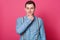 Peaceful thoughtful young boy stands wearing blue shirt, bow tie and stripped suspenders, posing isolated over pink background in