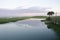 A peaceful, tranquil scene of a mirror lake with reeds, grass, trees, a street lamp, and a distant hill.