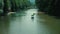 Pedal-powered catamarans float down the river. Dense green trees grow along the river banks.