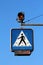 Pedestrians crossing blue and white road sign suspended from metal pole with two blinking yellow lights mounted on top