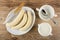 Peeled bananas, knife in dish, pitcher with yogurt, spoon in bowl on wooden table. Top view
